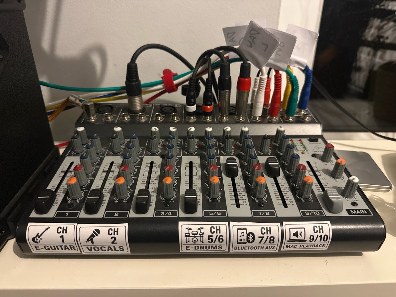 Sharp close-up of the labelled Behringer mixer, showing dedicated channels for electric guitar, vocals, electronic drums, Bluetooth aux and Mac playback.